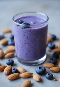A rich blue antioxidant protein smoothie in a glass, surrounded by fresh blueberries and almonds