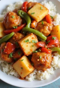 Healthy Baked Sweet and Sour Chicken A close-up of a plate of baked sweet and sour chicken served over white rice, featuring golden brown chicken pieces, cubed pineapples, green bell pepper strips, and red bell pepper chunks in a glossy sauce, garnished with chopped herbs