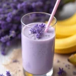 A tall glass of creamy lavender banana smoothie with a soft purple hue, garnished with fresh lavender blossoms and served with a pastel pink straw. The drink sits on a rustic beige cloth over a white wooden table, surrounded by ripe yellow bananas and sprigs of lavender in the background, creating a calm, aromatic, and refreshing scene. Made with banana, lavender syrup, and a sweetener.